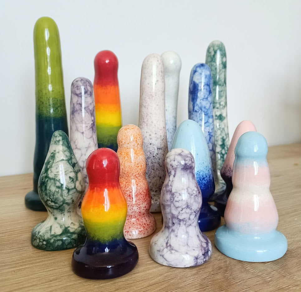 A group of various ceramic sex toys stand on a wooden table against a white background. The sex toys are brightly coloured with fun patterns, including rainbow, tie-dye, speckled and gradient patterns.