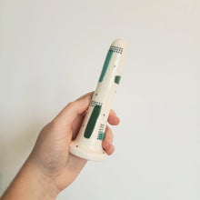 Load image into Gallery viewer, A hand holds a 5 inch ceramic dildo against a white background. The patterns on the dildo are teal and forest green strokes, with sets of dark green dots over-layed in sections, with more dots scattered across the white base colour.