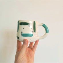 Load image into Gallery viewer, A hand holds a handmade ceramic mug against a white background. The patterns on the dildo are teal and forest green strokes, with sets of dark green dots over-layed in sections, with more dots scattered across the white base colour.
