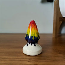 Load image into Gallery viewer, A 3 inch medium size cone shaped butt plug in a rainbow drip pattern flowing to a white base, stands on a wooden table