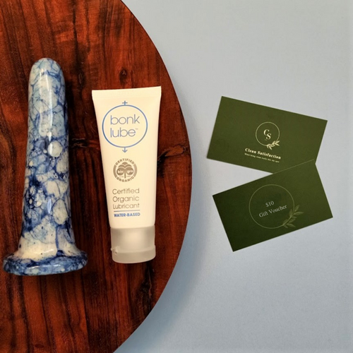 A 5 inch classic ceramic dildo in a dark blue and light blue bubble pattern lays on a round wooden board beside a tube of organic lubricant. The board is placed on a pale blue surface, which has two dark green business cards placed on it. One of the business cards has the words '$10 Gift Voucher' printed on it, while the other is the standard Clean Satisfaction business card.