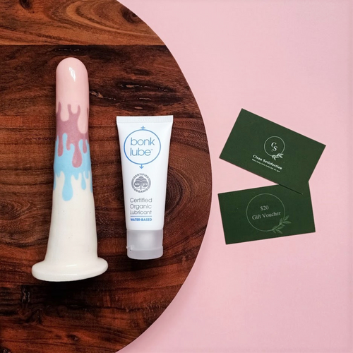 A 6 inch classic ceramic dildo in a pastel pink, purple and blue drip pattern lays on a round wooden board beside a tube of organic lubricant. The board is placed on a pale pink surface, which has two dark green business cards placed on it. One of the business cards has the words '$20 Gift Voucher' printed on it, while the other is the standard Clean Satisfaction business card.