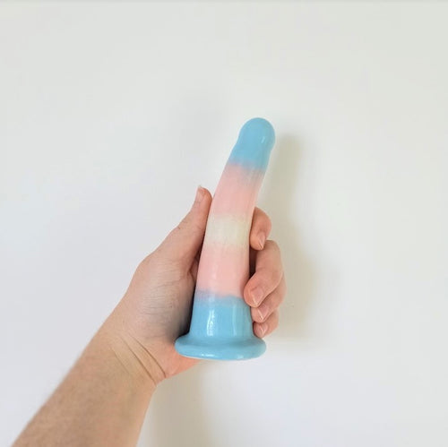 A hand holds a 6 inch curved ceramic dildo in a trans pride gradient pattern in front of a white background.