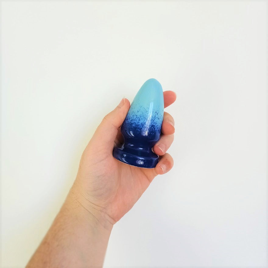 A hand holds a 3 inch cone shaped ceramic butt plug in a dark blue to light blue gradient pattern in front of a white background.