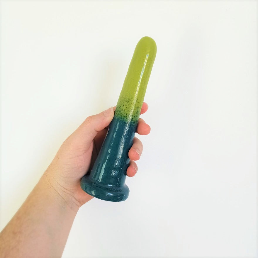 A hand holds a 7 inch classic ceramic dildo in a dark green to light green gradient pattern in front of a white background.