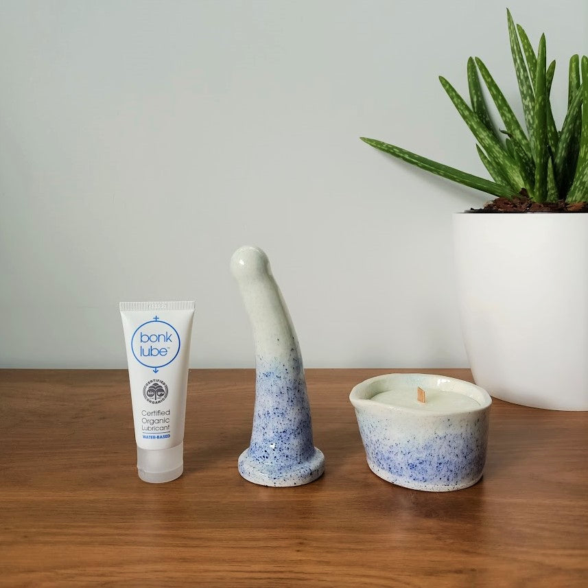 A tube of Bonk organic water-based lubricant, a 6 inch curved ceramic dildo and a matching massage candle in a dark blue to light blue speckle pattern stand in a row on a wooden table. An aloe vera plant in a white pot is visible to the right against a white background.
