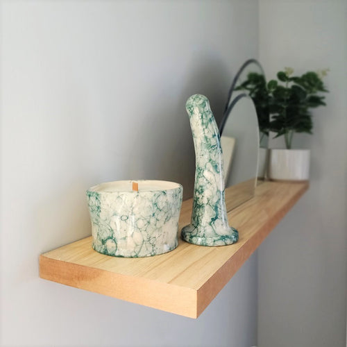 A handmade ceramic massage candle and a 6 inch curved ceramic dildo in a dark green bubble pattern stand at the end of a wooden shelf on a light grey wall. Two circular mirrors are inset into the shelf in the background, and a green plant in a white pot is visible at the other end of the shelf.