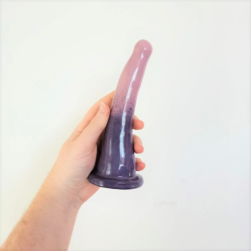A hand holds a 7 inch curved ceramic dildo in a dark purple to light purple gradient pattern in front of a white background.