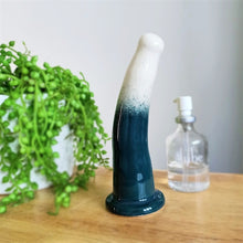 Load image into Gallery viewer, A 6 inch curved ceramic dildo in a white and dark green gradient pattern stands on a wooden table. A glass bottle of Uberlube stands to the right in the background, while a bright green plant flows from a white pot to the left.
