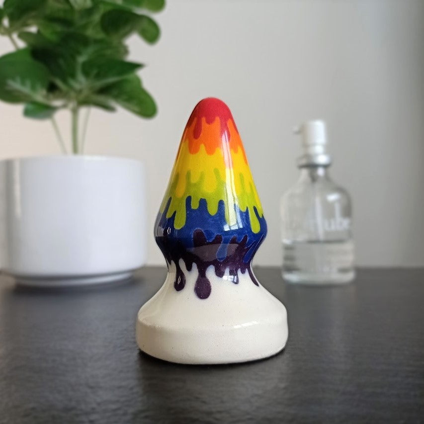 A 3 inch medium size cone shaped butt plug in a rainbow drip pattern flowing to a white base, stands on a black table. A plant in a white pot and a glass bottle of lubricant stand in the background.