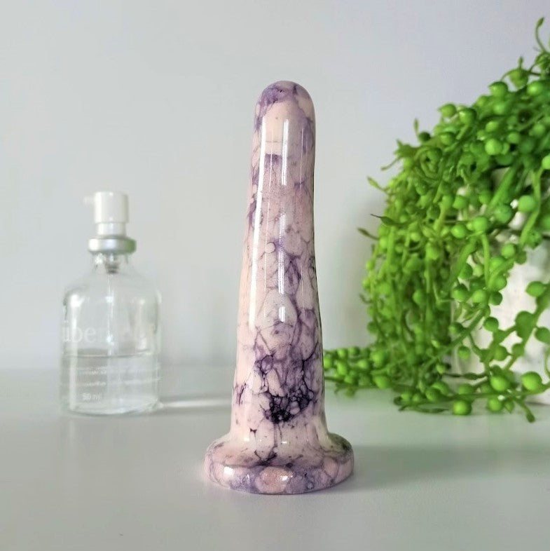 A 4 inch classic ceramic dildo in a pink and dark purple bubble pattern stands on a white table against a white background. A glass bottle of Uberlube is to the left in background, while a bright green pot plant flows from a white pot to the right.