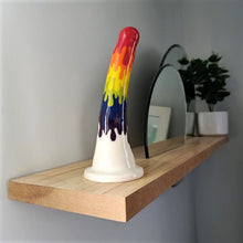 Load image into Gallery viewer, A 7 inch curved ceramic dildo in a rainbow drip pattern stands at the end of a wooden shelf. Two circular mirrors are inset into the shelf, and a dark green plant in a white pot stands at the other end of the shelf.

