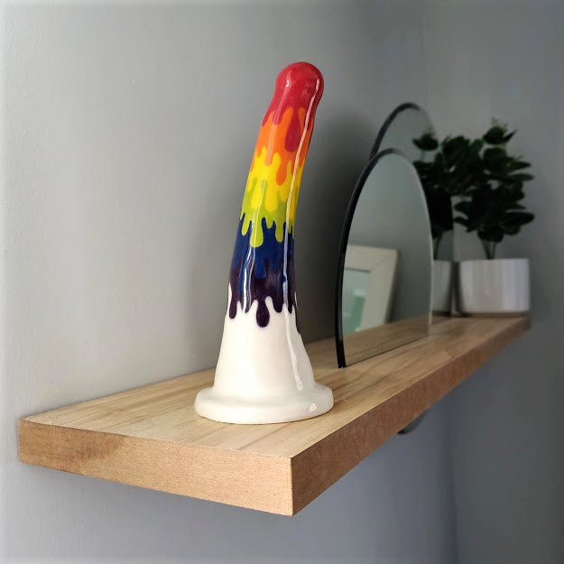 A 7 inch curved ceramic dildo in a rainbow drip pattern stands at the end of a wooden shelf. Two circular mirrors are inset into the shelf, and a dark green plant in a white pot stands at the other end of the shelf.