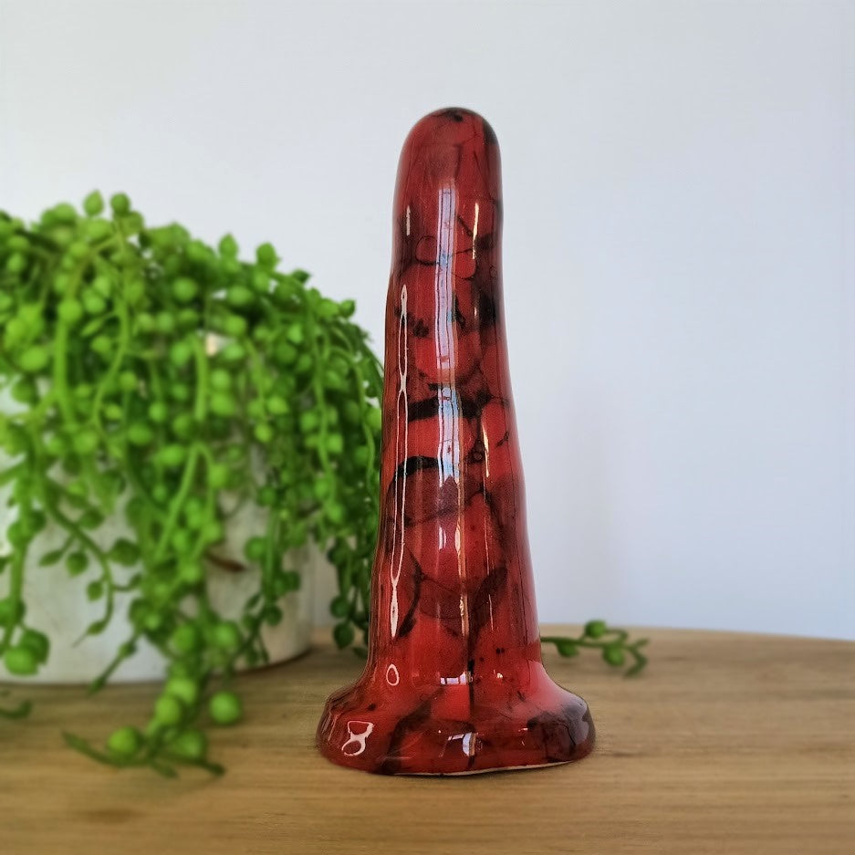 A 5 inch classic ceramic dildo in red with a black bubble pattern stands on a wooden table. A bright green plant flows from a white pot in the background.
