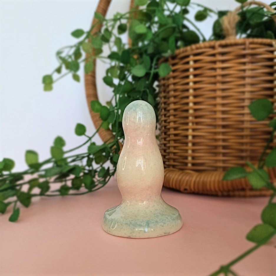 A 3 inch ceramic butt plug with 2 bumps in a trans pride speckle gradient pattern stands on a pink surface with a white background. A green plant flows from a wicker basket behind the butt plug.