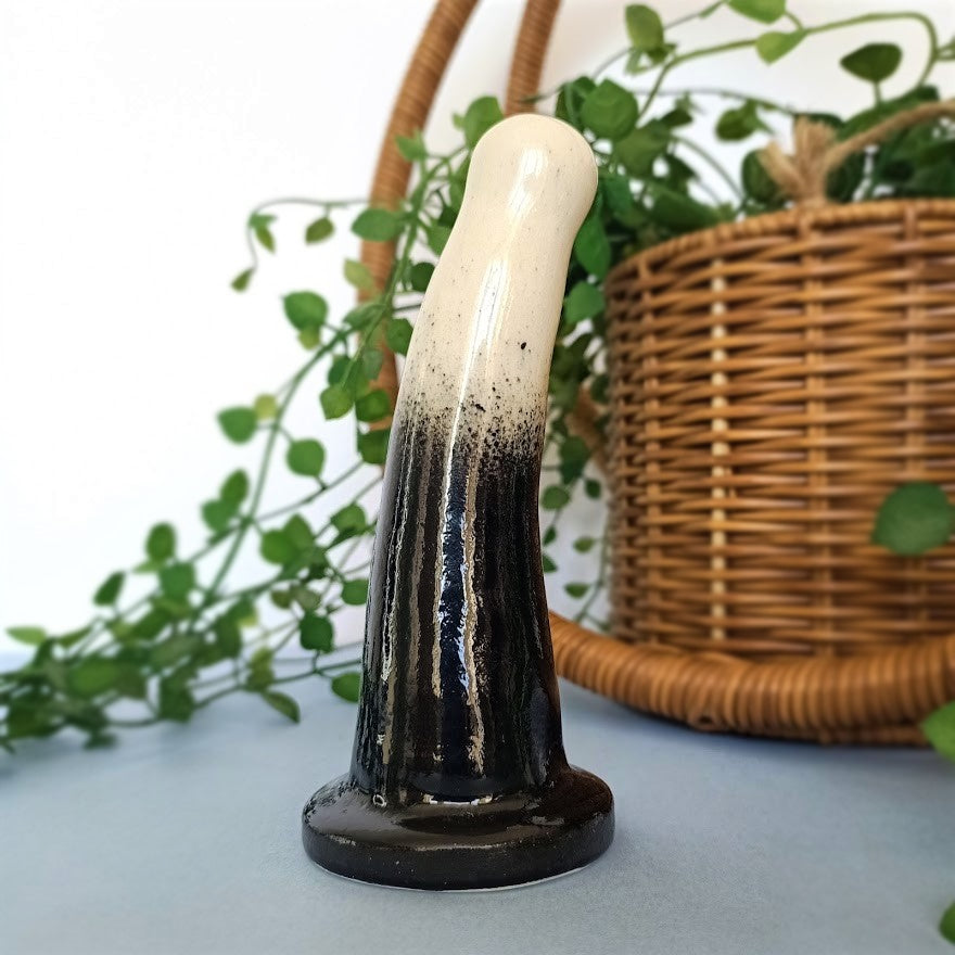 A 5 inch curved ceramic dildo in a white and black gradient pattern stands on a pale blue surface, against a white background. A pot plant flows behind the dildo from a pot in a wicker hanging basket.