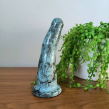 Load image into Gallery viewer, A 6 inch curved ceramic dildo in light blue with a black bubble pattern stands on a wooden surface with a white background. A bright green plant flows from a white pot in the background.
