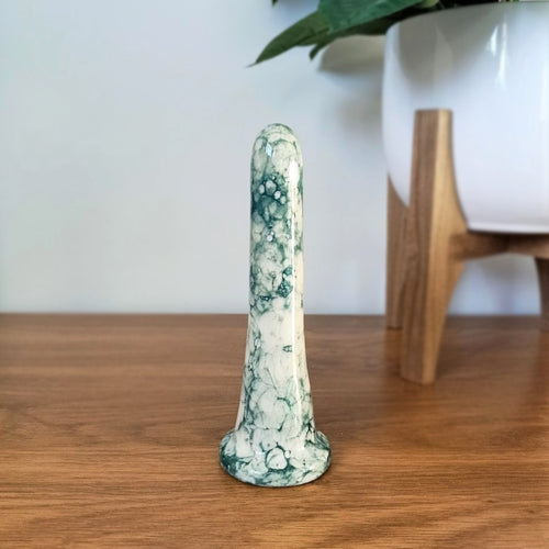 A 6 inch classic ceramic dildo in a dark green bubble pattern stands on a wooden table against a white background. A plant in a white pot with a wooden stand is to the right.