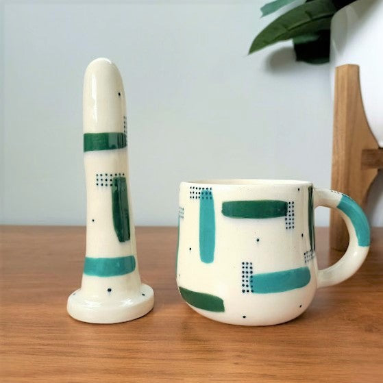A 5 inch classic ceramic dildo and a matching mug stand on a wooden table. The patterns on the products are teal and forest green strokes, with sets of dark green dots over-layed in sections, with more dots scattered across the white base colour.